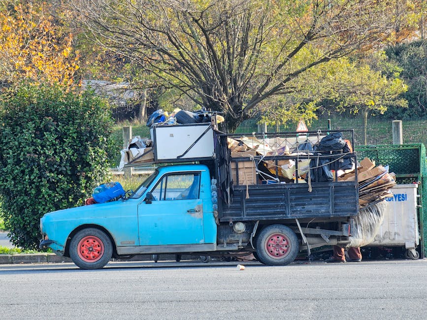 A blue, older-model pickup truck with a black metal cargo bed parked on the side of a street during daylight. The vehicle is loaded with various packing materials, cardboard boxes, plastic bags, and miscellaneous household items, all secured with ropes and straps. Some items, including a large white appliance or furniture piece wrapped in protective material, are positioned on top of the cargo area. Inside and around the cargo area, there are additional boxes and bags. The truck's wheels are pink, and the truck appears to be in the process of a home relocation or furniture transport, typical of an internal moving or clearance operation. In the background, a large tree with fall foliage and a green hedge are visible, with outdoor lighting indicating a bright day. The scene reflects the logistics involved in packing and moving household belongings, with the branding of Man with Van Leytonstone subtly present, aligning with house removals and moving services.