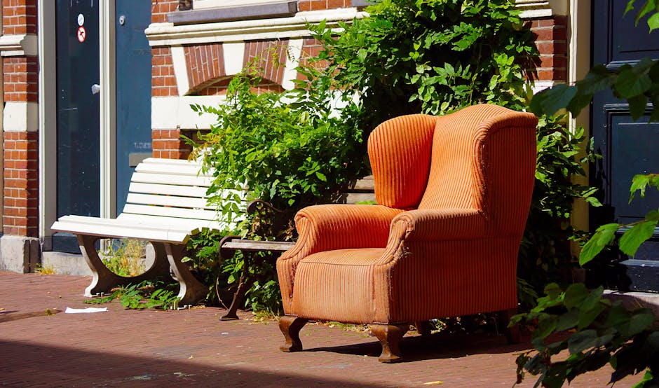 An outdoor scene on a brick-paved sidewalk in Leytonstone features an upholstered armchair with orange and red striped fabric, positioned next to a white wooden bench with decorative curved legs and slats for seating. The armchair is placed near a bush with green leaves, partially obscuring a black door on a red-brick building with white accents, suggesting it is before or after a home relocation or furniture transport process. Nearby, there are cardboard boxes and packing materials on the ground, indicating packing and moving activities. The lighting is bright, with shadows cast on the pavement, emphasizing the outdoor setting. This image, associated with Man with Van Leytonstone's removals service, illustrates the various furniture pieces and packing materials involved in house removals and furniture transport in Leytonstone.