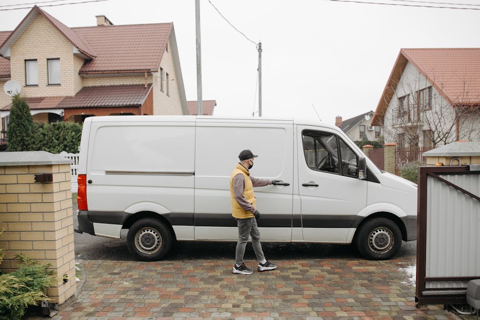 A man wearing a dark jacket and cap is loading cardboard boxes into the rear compartment of a large white moving van parked outside a modern industrial building with glass windows. Several packed card