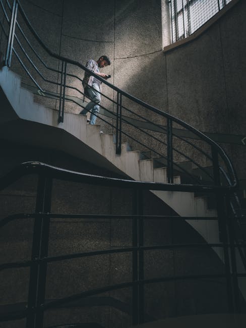 A person wearing a grey hoodie and dark trousers is walking up a curved concrete staircase inside a building, carrying a cardboard box wrapped in plastic for protection, which appears to contain household items. The staircase has black metal railings on both sides, with a textured, grey wall forming the background. The lighting is coming from a window on the right side, casting natural light onto the stairs and the individual. This scene likely depicts a home relocation or moving process, fitting into the context of professional removals services offered by Man with Van Leytonstone, as part of their house moving or furniture transport operations inside E11 flats. The environment shows a typical internal stairway used during packing and loading phases of a house removal, emphasizing careful handling and strategic navigation through staircases during furniture transport.