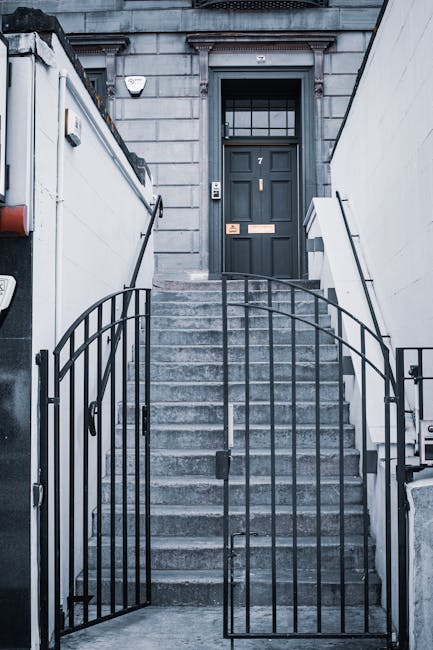 A person wearing a grey hoodie and dark trousers is walking up a curved concrete staircase inside a building, carrying a cardboard box wrapped in plastic for protection, which appears to contain household items. The staircase has black metal railings on both sides, with a textured, grey wall forming the background. The lighting is coming from a window on the right side, casting natural light onto the stairs and the individual. This scene likely depicts a home relocation or moving process, fitting into the context of professional removals services offered by Man with Van Leytonstone, as part of their house moving or furniture transport operations inside E11 flats. The environment shows a typical internal stairway used during packing and loading phases of a house removal, emphasizing careful handling and strategic navigation through staircases during furniture transport.