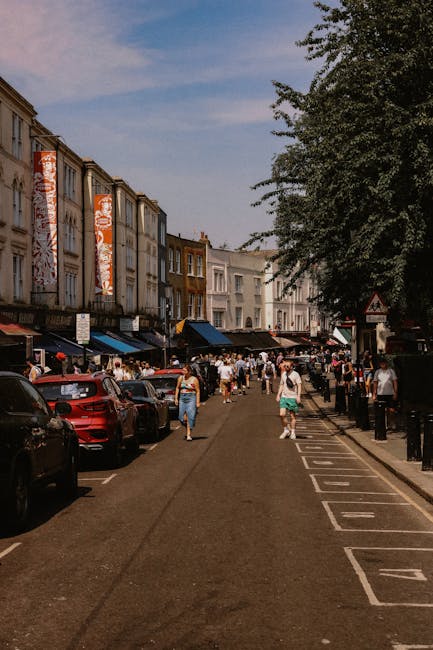 A man wearing a red cap, red t-shirt, and navy trousers is standing on the pavement next to an open white van, which is parked on a residential street surrounded by green trees. He is in the process o