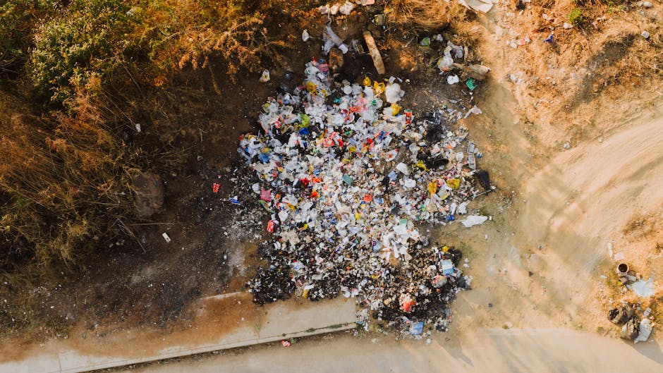 Aerial view of a large pile of mixed household waste and recyclable materials, including plastic bottles, cardboard, and paper, situated in an open dirt area adjacent to a paved road. The surrounding environment features dry, sparse vegetation along the edges of the waste pile, indicating a suburban or semi-rural location. It appears to be a waste disposal site or illegal dumping area, with no visible safety barriers or signage. The image highlights the need for proper waste management and recycling services, similar to those provided by Man with Van Leytonstone for responsible house removals and furniture transport, especially during home relocation and moving projects.