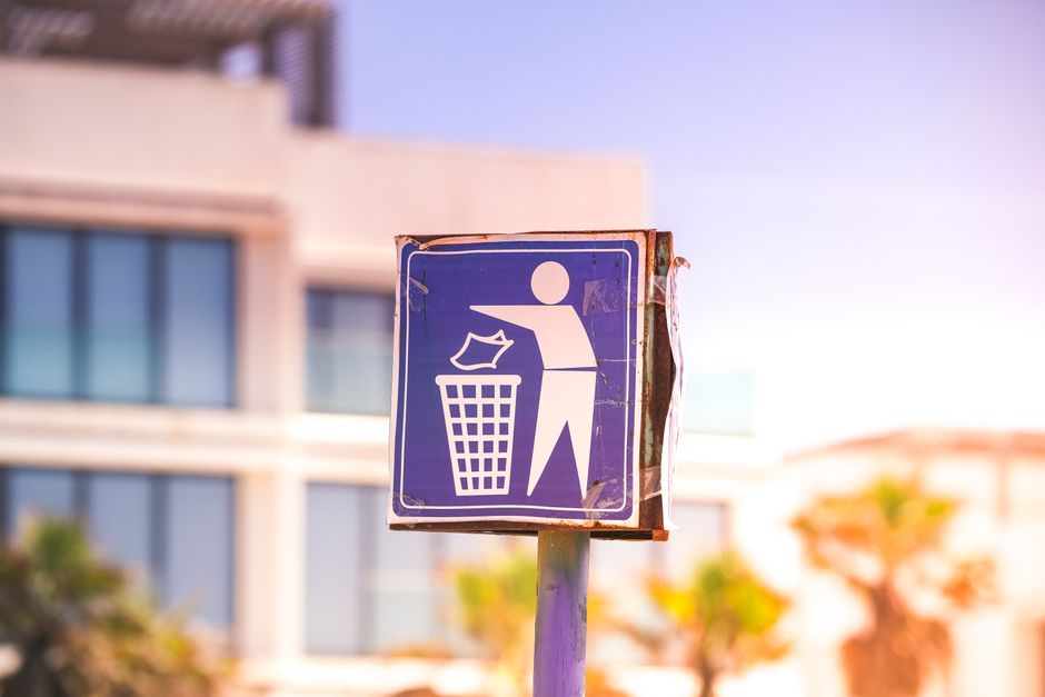 A close-up of a weathered, square blue public waste disposal sign mounted on a metal pole, featuring a white pictogram of a person discarding paper into a trash bin. The sign appears slightly damaged with visible scratches and peeling edges. In the background, there are modern residential buildings with large windows and balconies, and some blurred trees and sky, suggesting an urban setting during daytime. The scene may relate to house removals or relocation services by Man with Van Leytonstone, emphasizing waste disposal guidelines relevant to moving processes, including packing and clutter management as part of home relocation activities.
