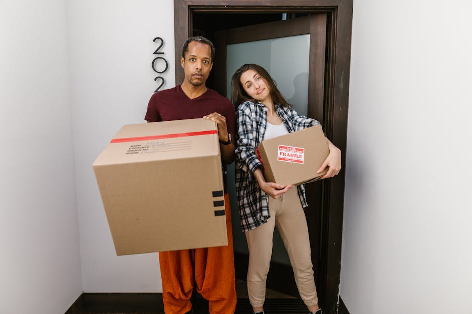 A man and a woman stand inside a doorway, each holding a cardboard moving box. The man, wearing an orange pair of pants and a maroon shirt, is holding a large box with black tape and facing forward. The woman, dressed in light-colored pants, a checkered shirt, and a white top, is holding a smaller box marked with a red and white 'Fragile' label, leaning slightly against the doorframe. The interior background includes a plain wall with the apartment number '2002' visible on the left side, and the doorway has a dark wooden frame. The scene depicts a home relocation process, with the individuals preparing for furniture transport and packing and moving activities, likely as part of a professional removals service by Man with Van Leytonstone. The lighting is natural and evenly distributed, emphasizing the packing materials and the loading process associated with house removals and moves.