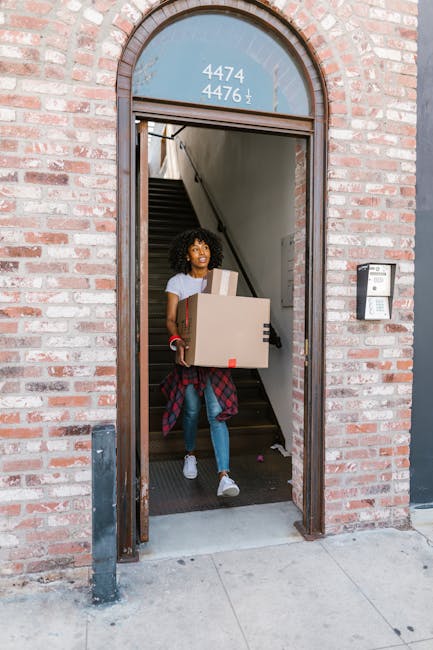 A young woman with curly hair, wearing a white t-shirt, blue jeans, and white sneakers, is standing at the entrance of a brick building, holding three cardboard boxes of varying sizes. She is positioned just inside the doorway, which leads to a stairwell with dark treads visible behind her. The doorway has a rounded top with house number signage above, displaying the numbers '4474', '4476', and an arrow pointing downward. The building exterior features a red-brick façade with a metal mailbox affixed to the right side of the doorframe. The woman appears to be in the process of moving or home relocation, with a plaid shirt tied around her waist, and is surrounded by packing materials, suggesting a packing and moving process. An intercom or access control panel is mounted on the wall to her right. This scene depicts the routine of a professional house removal or furniture transport service, such as offered by Man with Van Leytonstone, and aligns with the themes of moving logistics and packing for relocations.