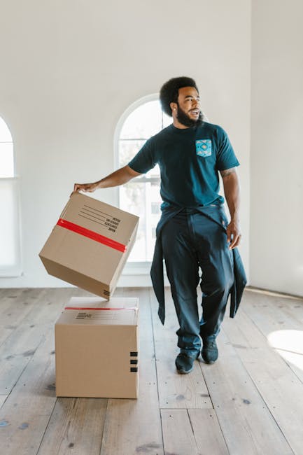 A man with a beard and curly hair dressed in a navy blue t-shirt and dark trousers is inside a bright, empty room with white walls and large arched windows, carrying a cardboard box with red tape. The