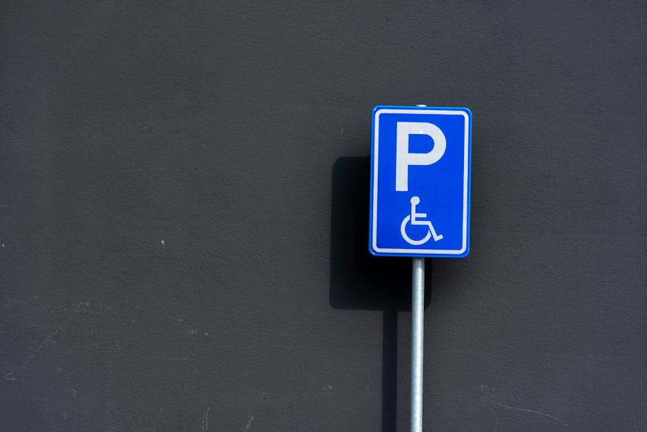 A close-up image of a blue parking permit sign with a white 'P' and a wheelchair symbol, mounted on a metal pole against a dark grey exterior wall. The sign indicates designated parking for disabled persons and is located in an outdoor setting, possibly near a property or loading area. The shadow of the sign can be seen cast on the wall, suggesting natural lighting. The image is relevant to house removals and relocation services, as it depicts parking permissions that are often necessary for professional moving companies like Man with Van Leytonstone when executing home relocation, furniture transport, or packing and moving tasks in the Leytonstone area, specifically in Waltham Forest.